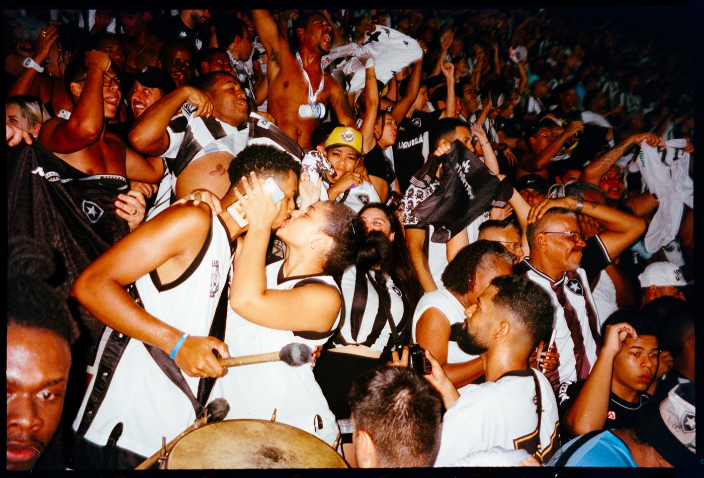Multidão de torcida em preto e branco, com um casal se beijando no meio e um surdo em primeiro plano