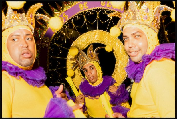 Três foliões de capuz amarelo e gola roxa, dois de frente para a câmera e um ao centro, emoldurado por um sol dourado